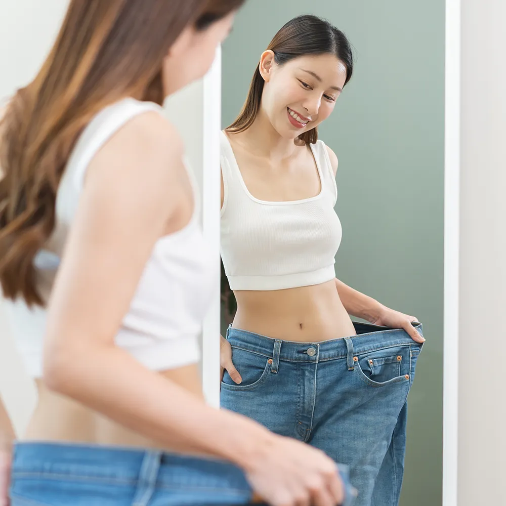 A woman happily looking in the mirror, noticing her pants fit looser after weight loss.