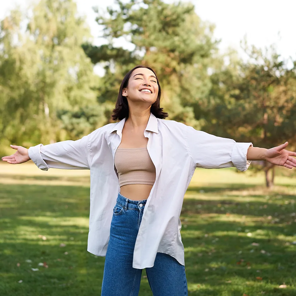 A woman in a park with open arms, smiling with closed eyes, enjoying the moment.