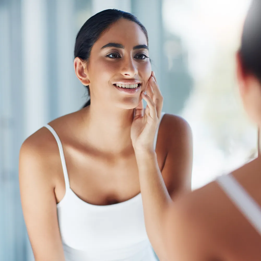 A woman happily looking at her reflection in the mirror while touching her skin.